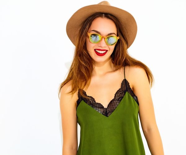 Young stylish girl model in casual summer green clothes and brown hat with red lips, posing near white wall